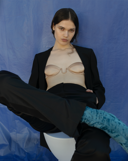 A model in a black blazer and pants sits on a white stool in front of a bright blue backdrop, posing in a bra top with patterned blue hosiery.
