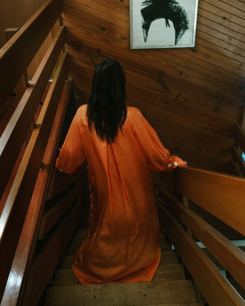 Back view of a woman with long black hair ascending wooden stairs in an orange long-sleeve dress, complemented by an abstract art piece.