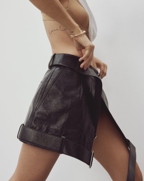 Fashion editorial shot of a woman wearing a black leather mini skirt with a large belt, captured in a studio setting against a white backdrop.
