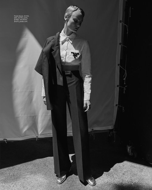 Editorial fashion shot of a slender adult female in a suit, standing in front of a white backdrop, photographed in black and white.