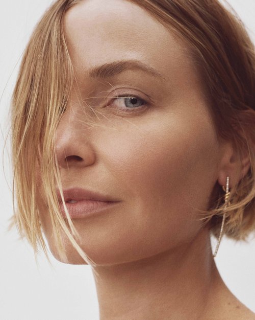 Close-up portrait of a fair-skinned adult woman with blonde hair parted to the side, wearing gold earrings, against a plain white background.