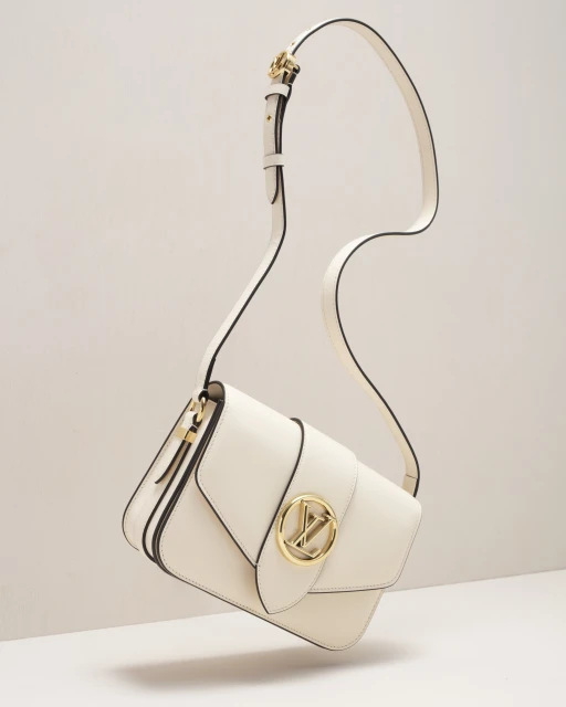 Studio shot of a cream-colored leather shoulder bag with a gold logo buckle, suspended by its strap against an off-white background.