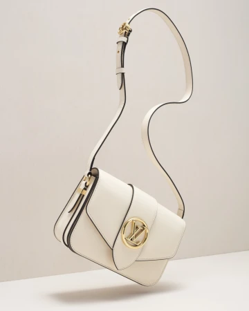 Studio shot of a cream-colored leather shoulder bag with a gold logo buckle, suspended by its strap against an off-white background.