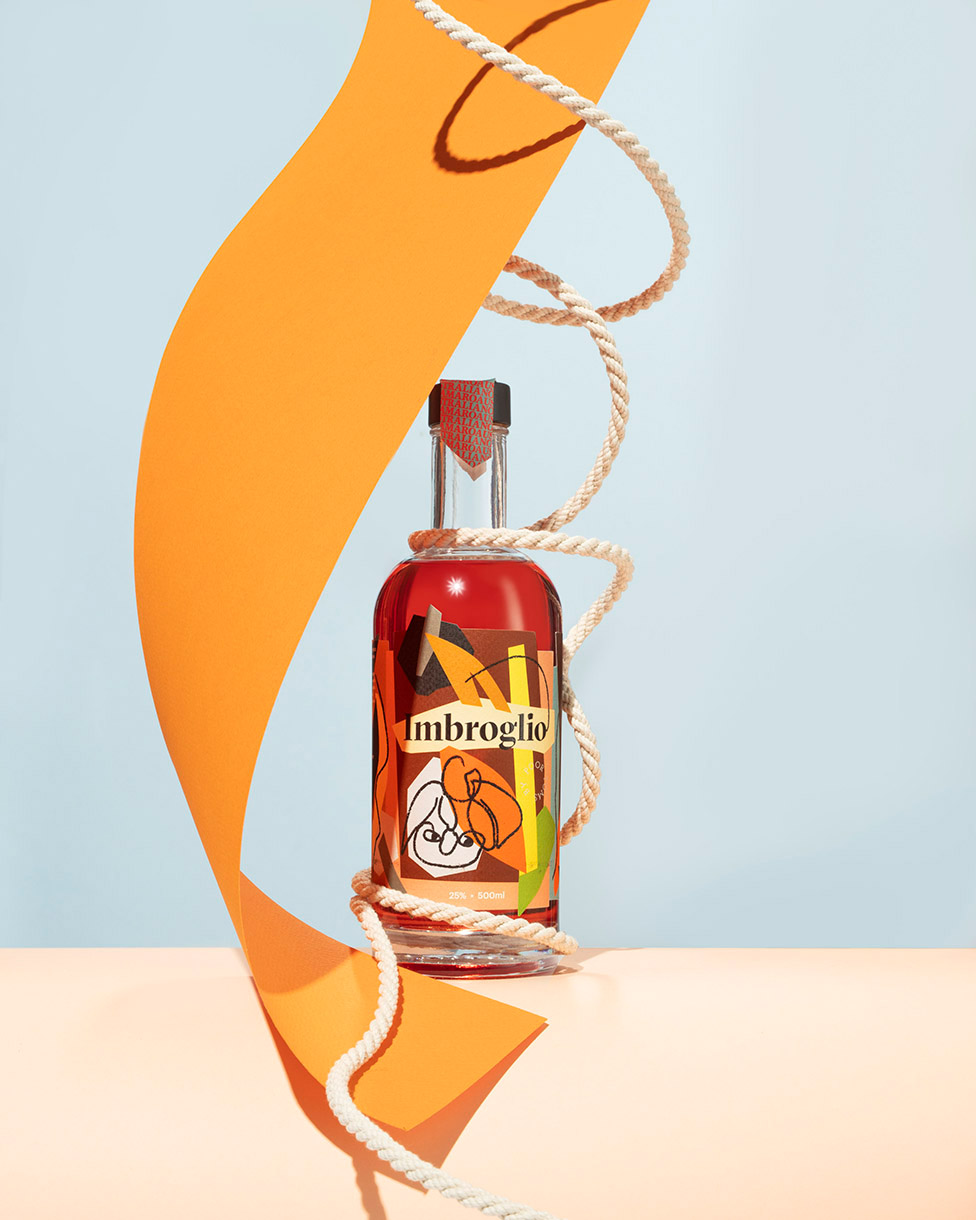 Product shot of Imbroglio bottle with orange paper shapes and rope on peach surface with blue background in a studio setup.