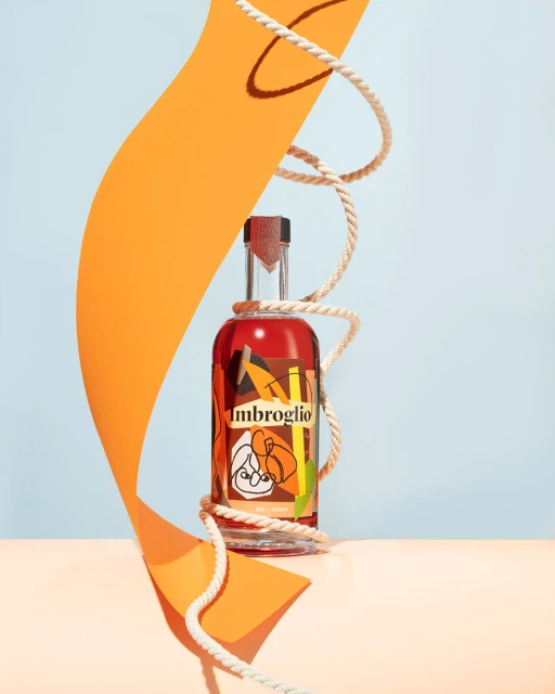 Product shot of Imbroglio bottle with orange paper shapes and rope on peach surface with blue background in a studio setup.