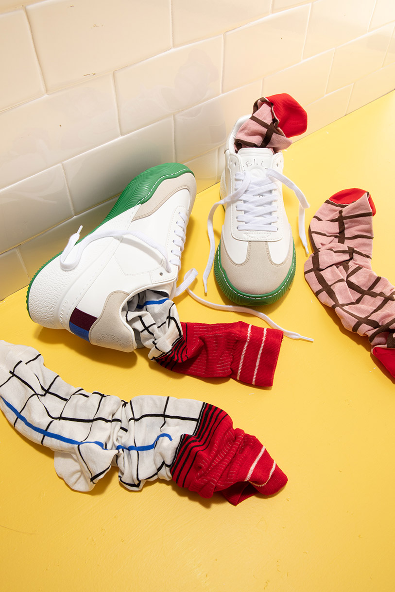 Still life of two white sneakers with colorful soles and laces, next to patterned socks, arranged on a yellow surface with tile background.