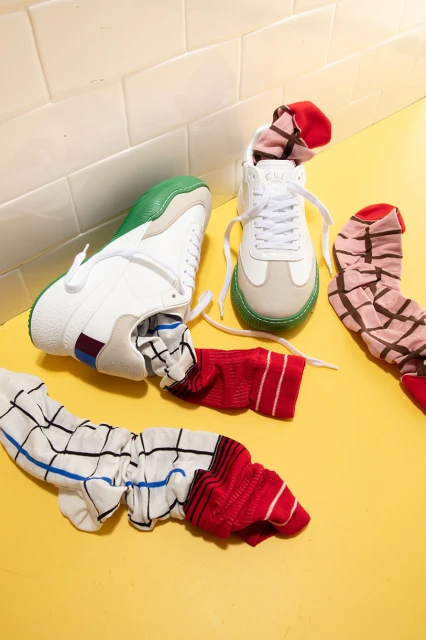 Still life of two white sneakers with colorful soles and laces, next to patterned socks, arranged on a yellow surface with tile background.
