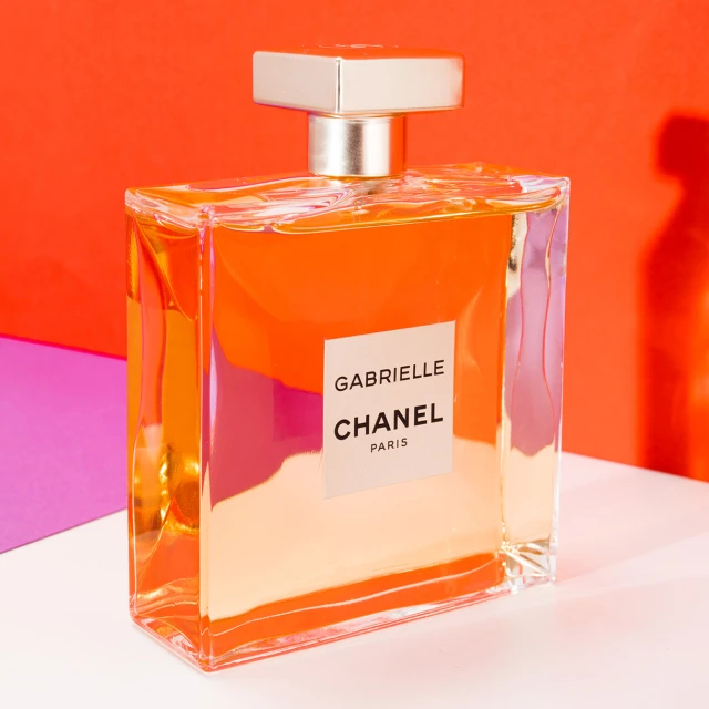 Elegant, square-shaped Chanel Gabrielle perfume bottle on a white surface against a vivid coral and fuchsia backdrop in studio lighting.