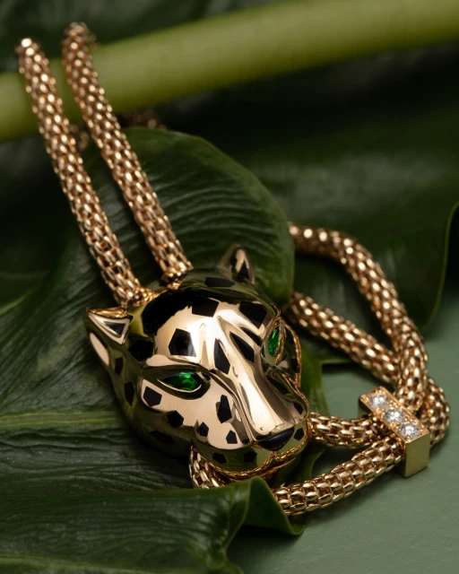 Gold tone necklace with jaguar pendant, featuring emerald-green eyes and black enamel spots, resting on textured green leaves.