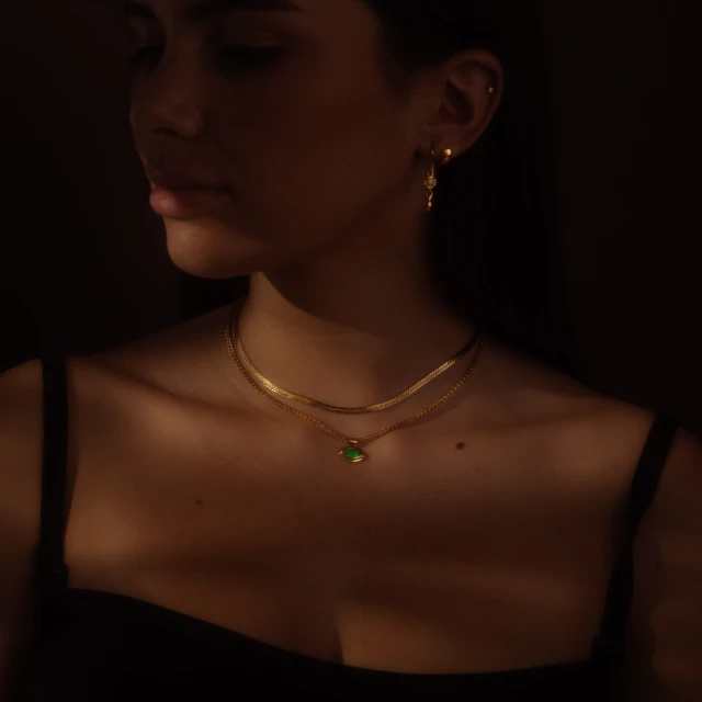 Young woman in studio light wearing a gold necklace with green gem, gold earrings, and a black top against a dark brown background.