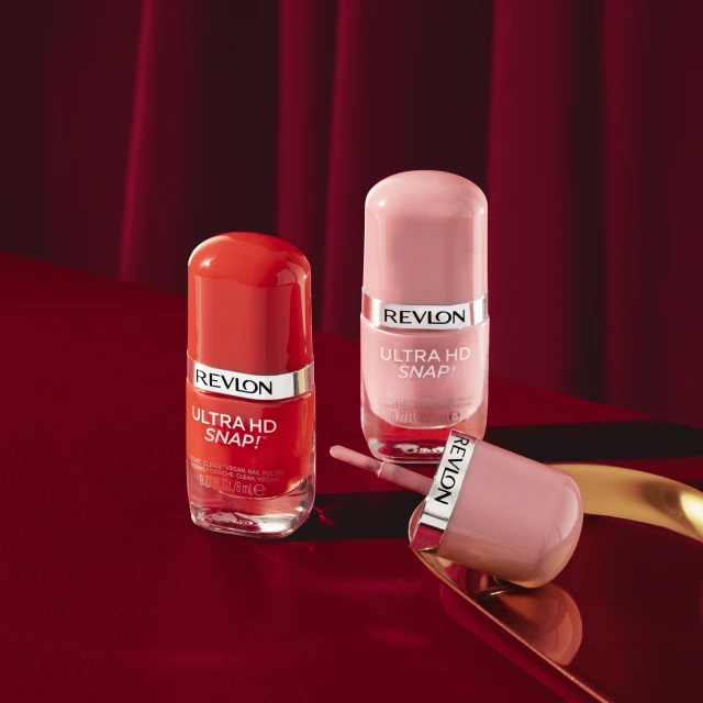 Revlon Ultra HD Snap! nail polish bottles in coral and light pink on a red surface with red draped fabric in the background.