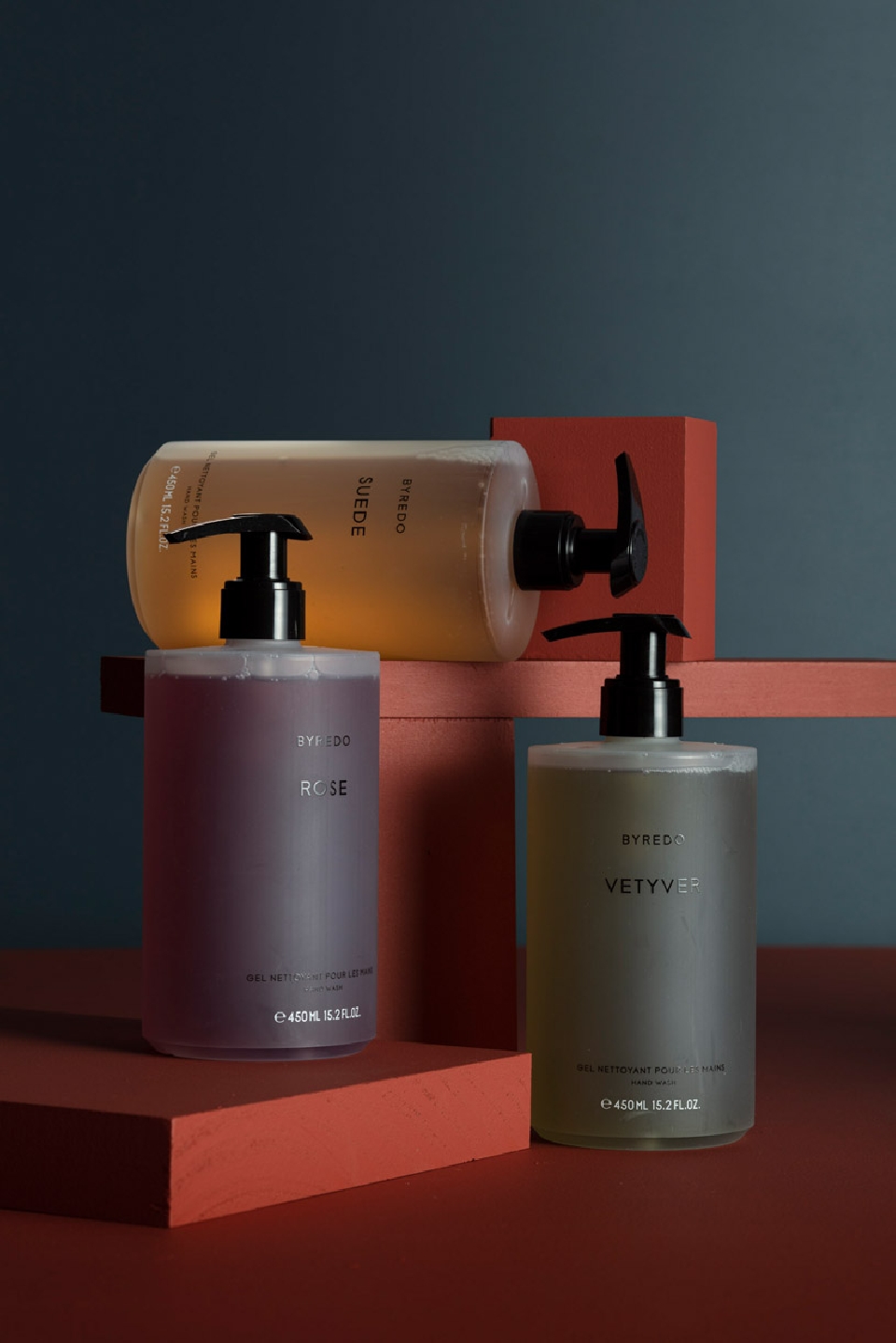 Three Byredo bottles are displayed on red cube platforms against a blue background, featuring a rose-scented bottle in the foreground.