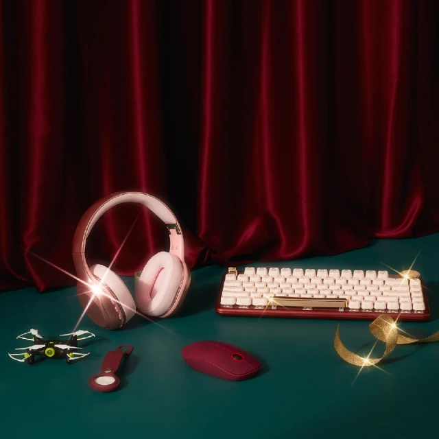 Gaming setup on a teal surface features a cream keyboard, pink headphones, burgundy mouse, drone, key chain, and gold ribbon against a red curtain.