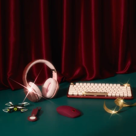 Gaming setup on a teal surface features a cream keyboard, pink headphones, burgundy mouse, drone, key chain, and gold ribbon against a red curtain.