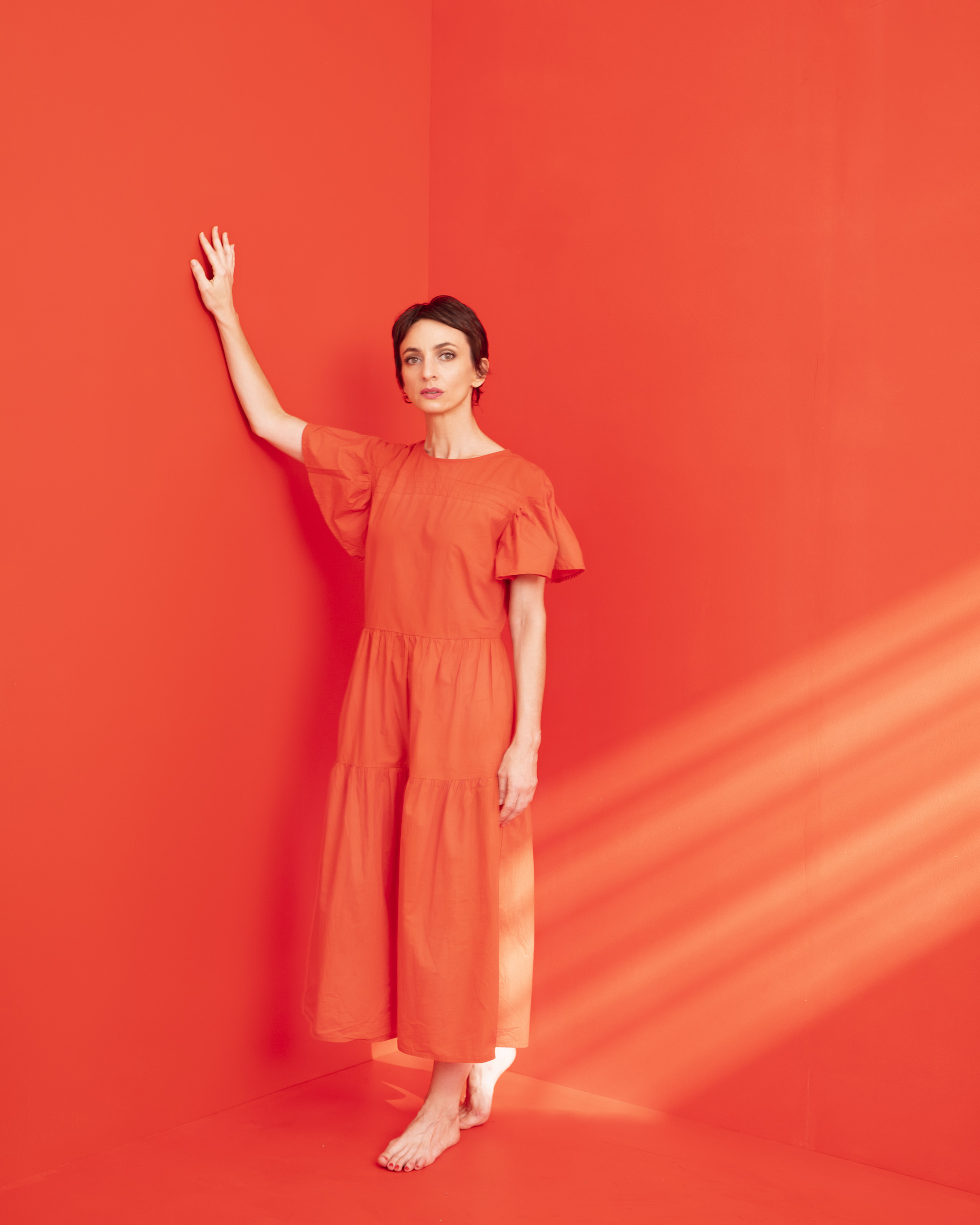 Full-length shot of an adult woman in an orange dress, striking a pose with her arm raised, against a vibrant orange wall indoors.