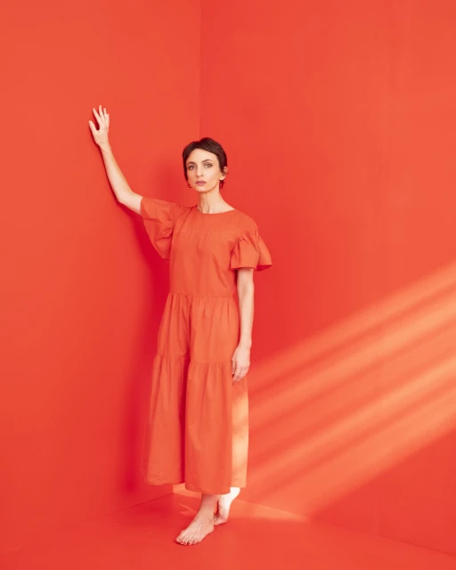 Full-length shot of an adult woman in an orange dress, striking a pose with her arm raised, against a vibrant orange wall indoors.
