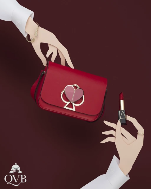 Red handbag displayed with lipstick, complemented by hands wearing white sleeves and gold jewelry, set against a solid burgundy backdrop.