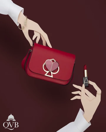 Red handbag displayed with lipstick, complemented by hands wearing white sleeves and gold jewelry, set against a solid burgundy backdrop.