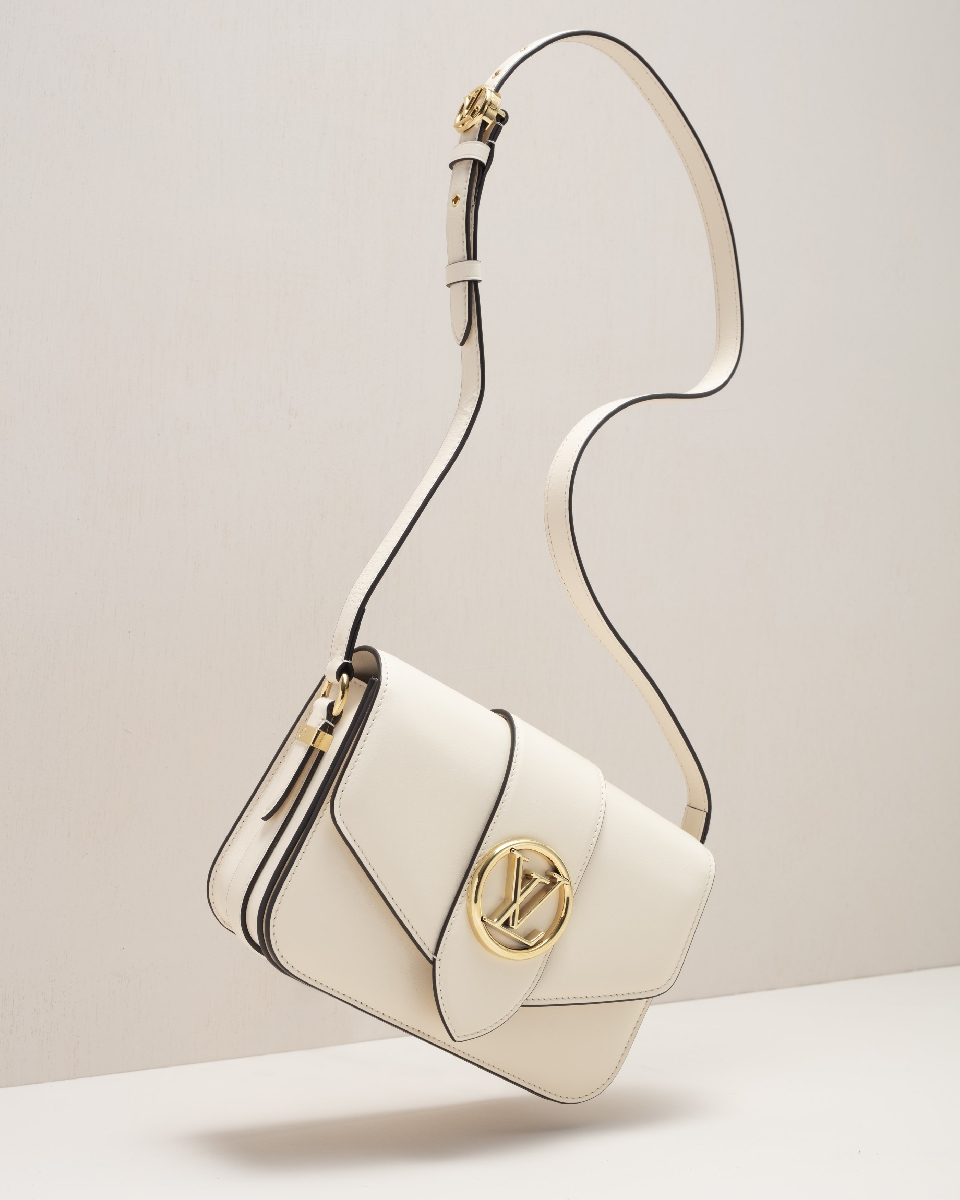 Elevated studio shot of a modern cream-colored crossbody bag with a gold logo and black piping. The bag is suspended in mid-air.