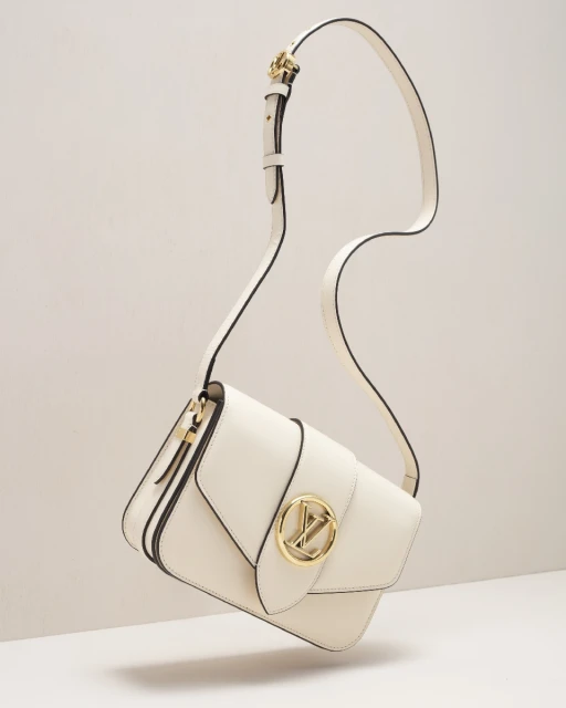 Elevated studio shot of a modern cream-colored crossbody bag with a gold logo and black piping. The bag is suspended in mid-air.