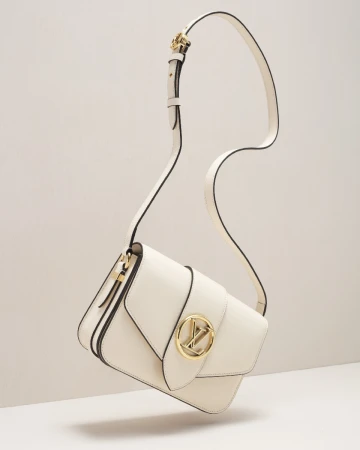 Elevated studio shot of a modern cream-colored crossbody bag with a gold logo and black piping. The bag is suspended in mid-air.