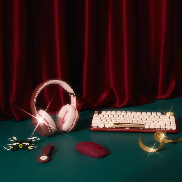Still life photo featuring pink headphones, white keyboard, burgundy mouse, drone, keychain, and gold ribbon against a red curtain backdrop.