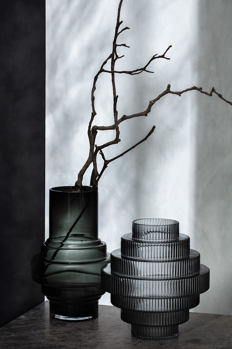 Still life of a gray textured vases in front of a mottled white and gray backdrop with a dark branch inside one vase.