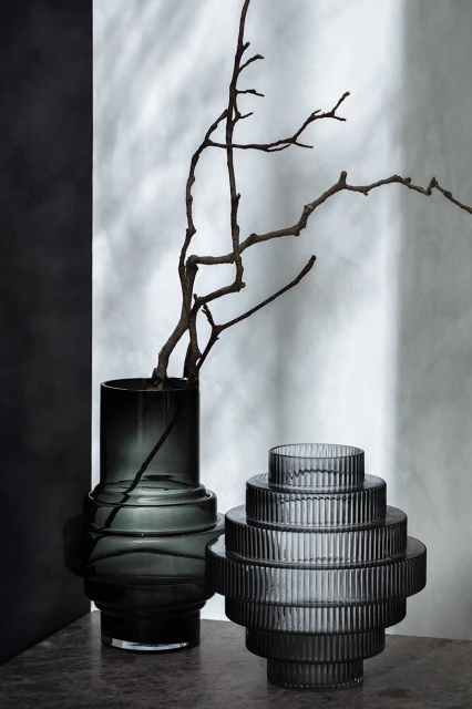 Still life of a gray textured vases in front of a mottled white and gray backdrop with a dark branch inside one vase.