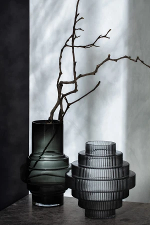 Still life of a gray textured vases in front of a mottled white and gray backdrop with a dark branch inside one vase.