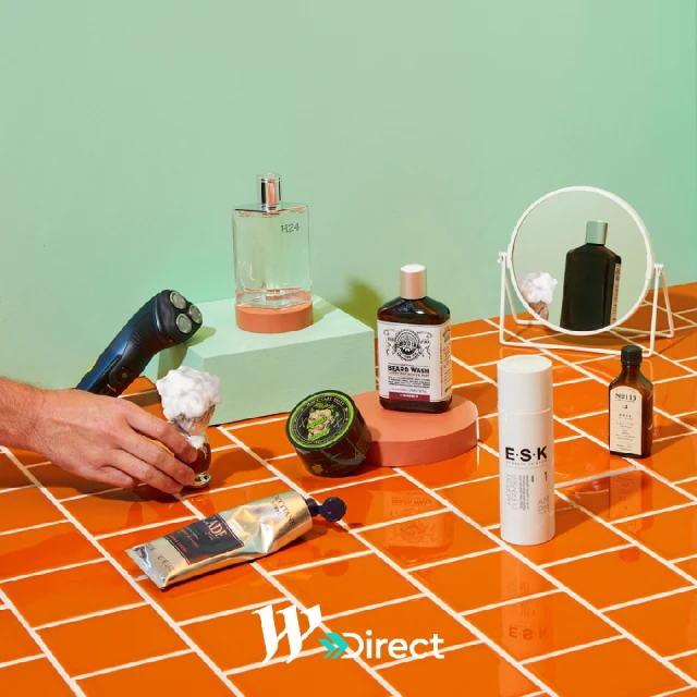 A diverse set of men's grooming products is displayed on orange tile against a split mint green and white background, studio lit.