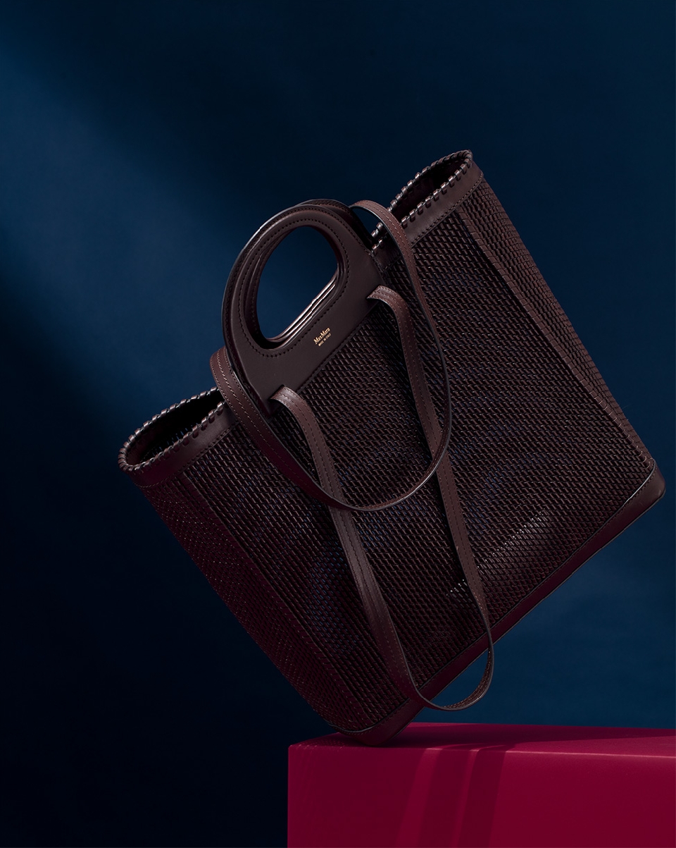 Elegant woven tote bag in brown leather, angled studio shot against a dark blue background with a magenta block.