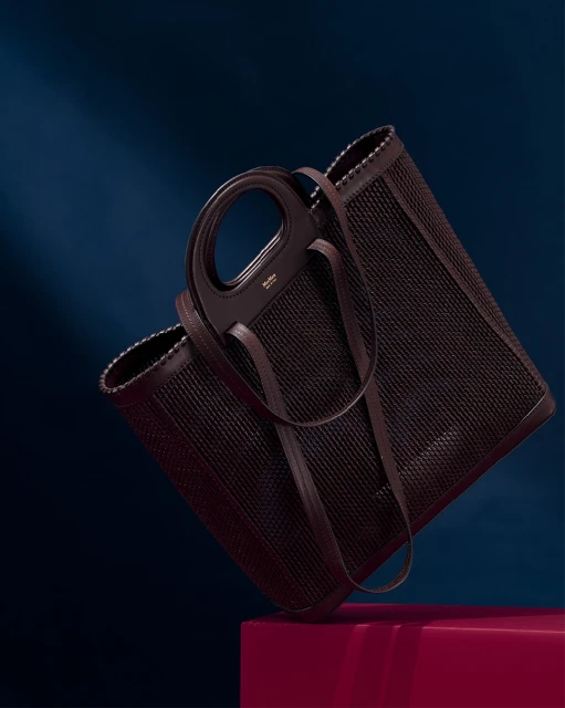 Elegant woven tote bag in brown leather, angled studio shot against a dark blue background with a magenta block.