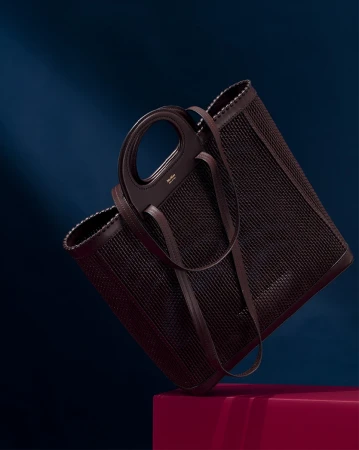 Elegant woven tote bag in brown leather, angled studio shot against a dark blue background with a magenta block.