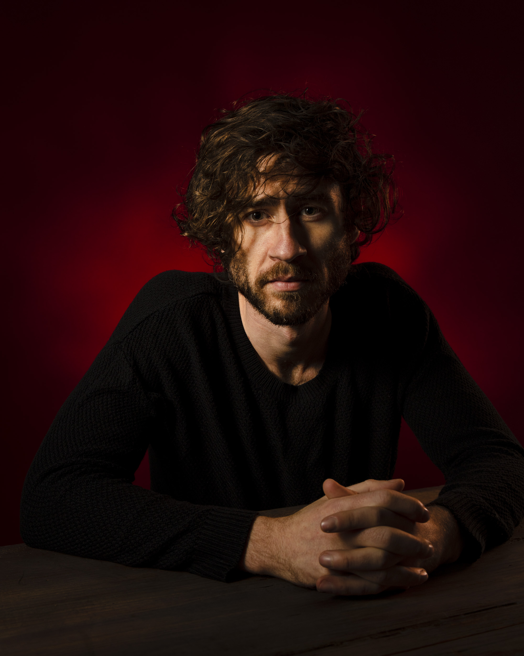 Portrait of a man with curly hair sitting at a table against a red backdrop. He wears a black long-sleeved shirt, with hands clasped.