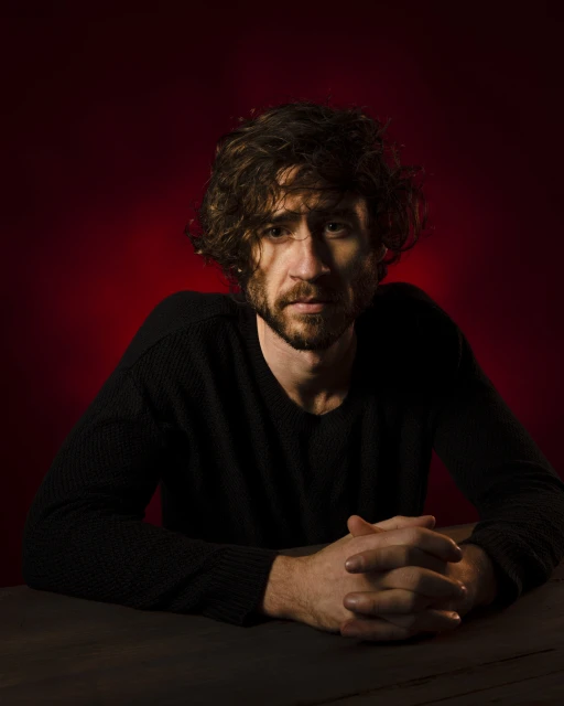Portrait of a man with curly hair sitting at a table against a red backdrop. He wears a black long-sleeved shirt, with hands clasped.