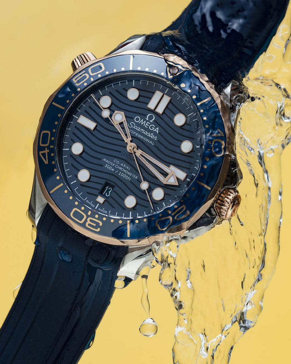 Omega Seamaster watch close-up: blue dial with gold accents, stainless steel case, rubber strap, water droplets, against a yellow backdrop.