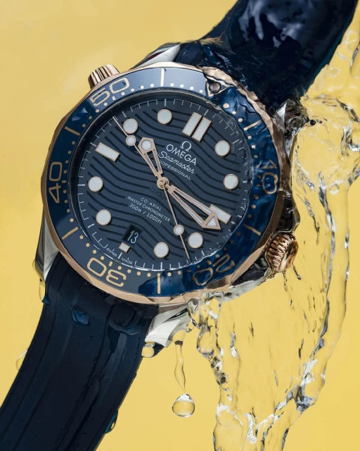 Omega Seamaster watch close-up: blue dial with gold accents, stainless steel case, rubber strap, water droplets, against a yellow backdrop.