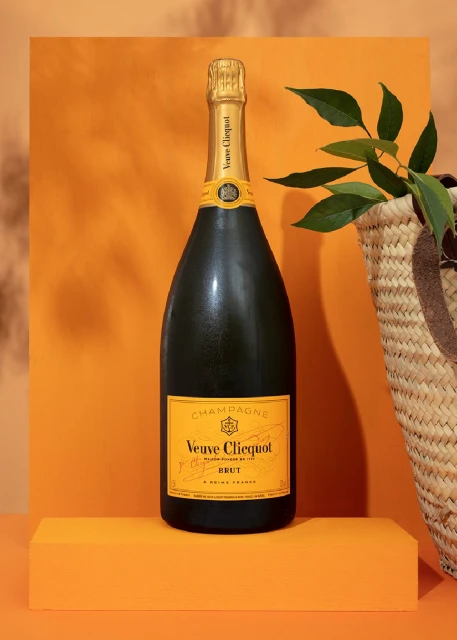 A bottle of Veuve Clicquot Champagne stands on an orange block with a straw bag and green leaves in the background.