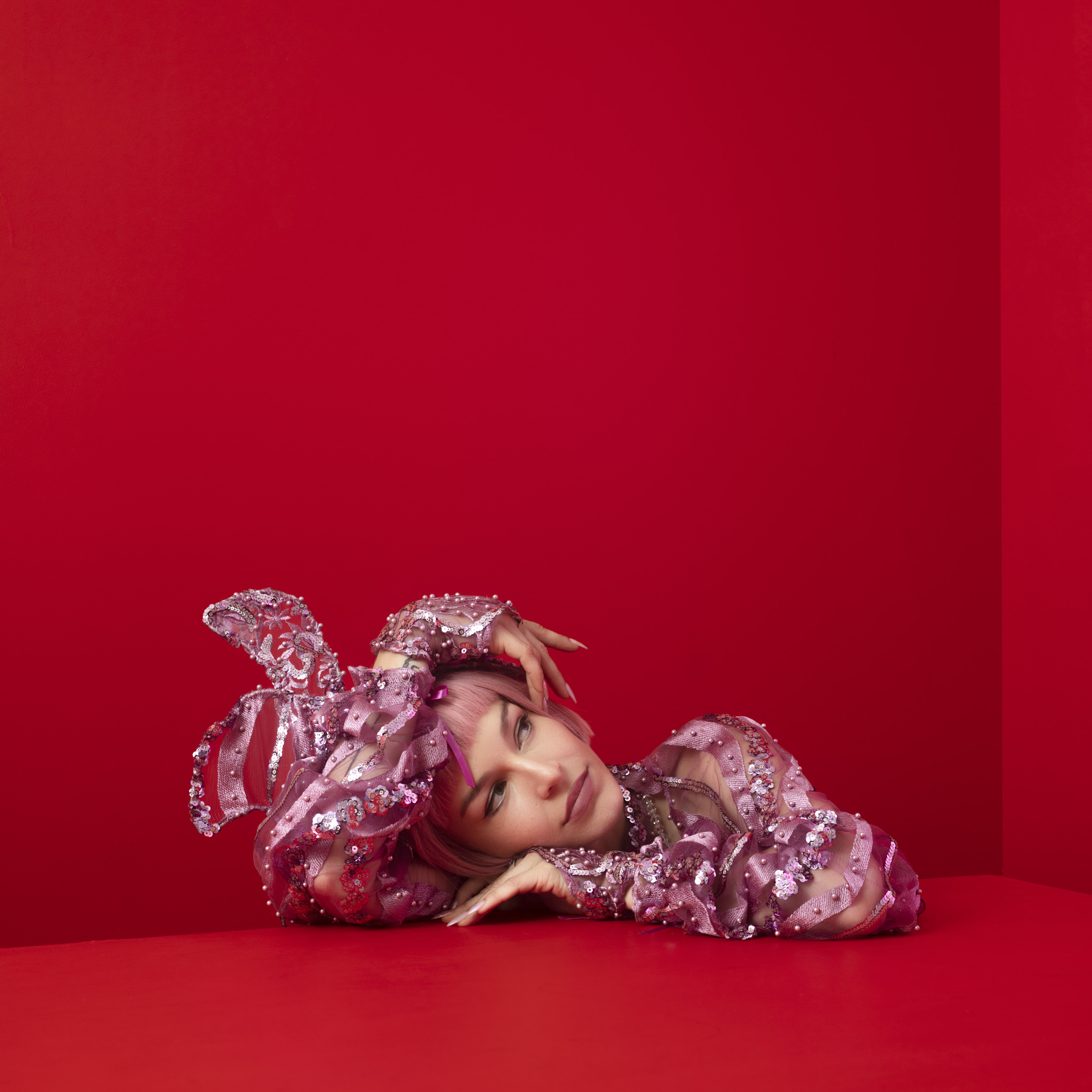 Young woman with pink hair in embellished pink top laying on a red surface in front of a red wall looking at the camera.