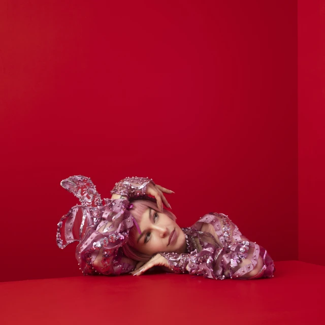 Young woman with pink hair in embellished pink top laying on a red surface in front of a red wall looking at the camera.