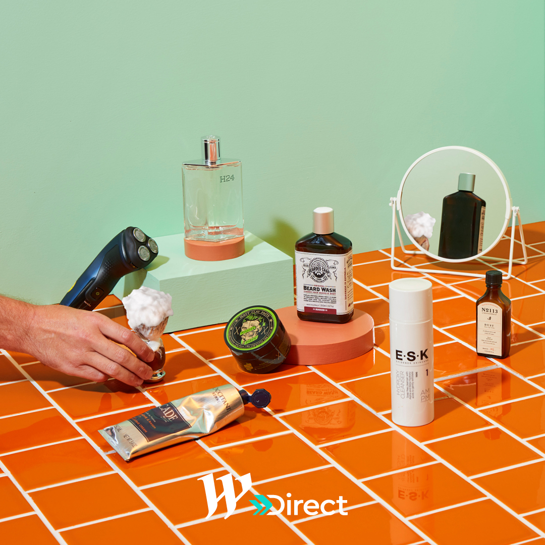 Several men's grooming products are displayed on an orange tiled surface in a bright photo studio setting with a mint green backdrop.