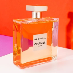 A rectangular glass perfume bottle with amber liquid sitting on a white surface against a bright orange and purple backdrop.