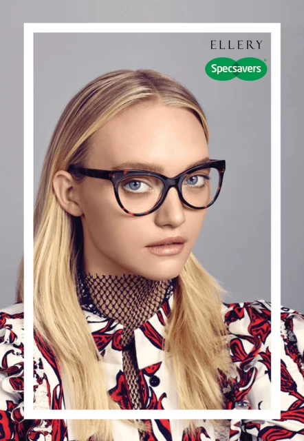 Blonde woman with fair skin and neutral expression poses in a white and red patterned shirt, wearing cat-eye glasses against a grey background.