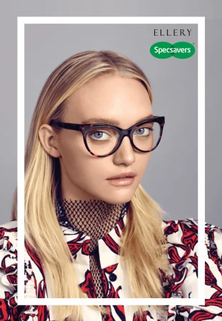 Blonde woman with fair skin and neutral expression poses in a white and red patterned shirt, wearing cat-eye glasses against a grey background.