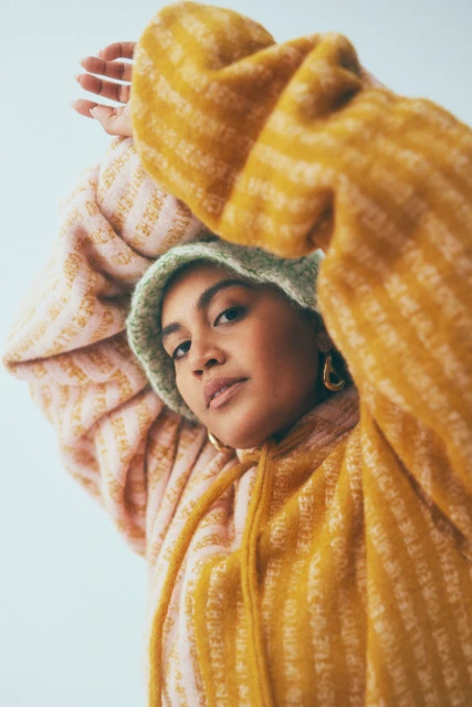 Portrait of a woman with a knit hat and gold earrings wearing a striped hoodie, posing with arms raised against a white background.