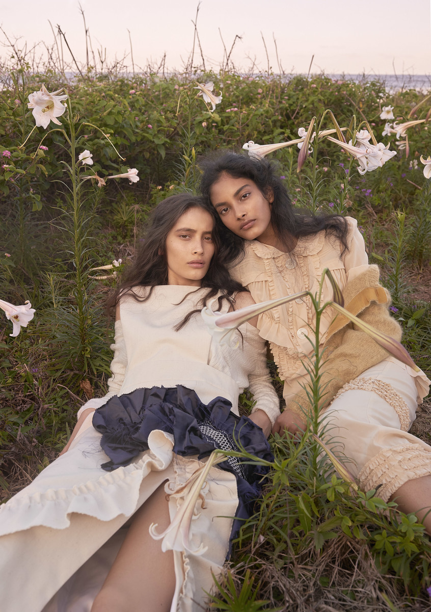 Two women lie in a lush field of white lilies, wearing elegant, ruffled cream and tan colored dresses in an outdoor setting.