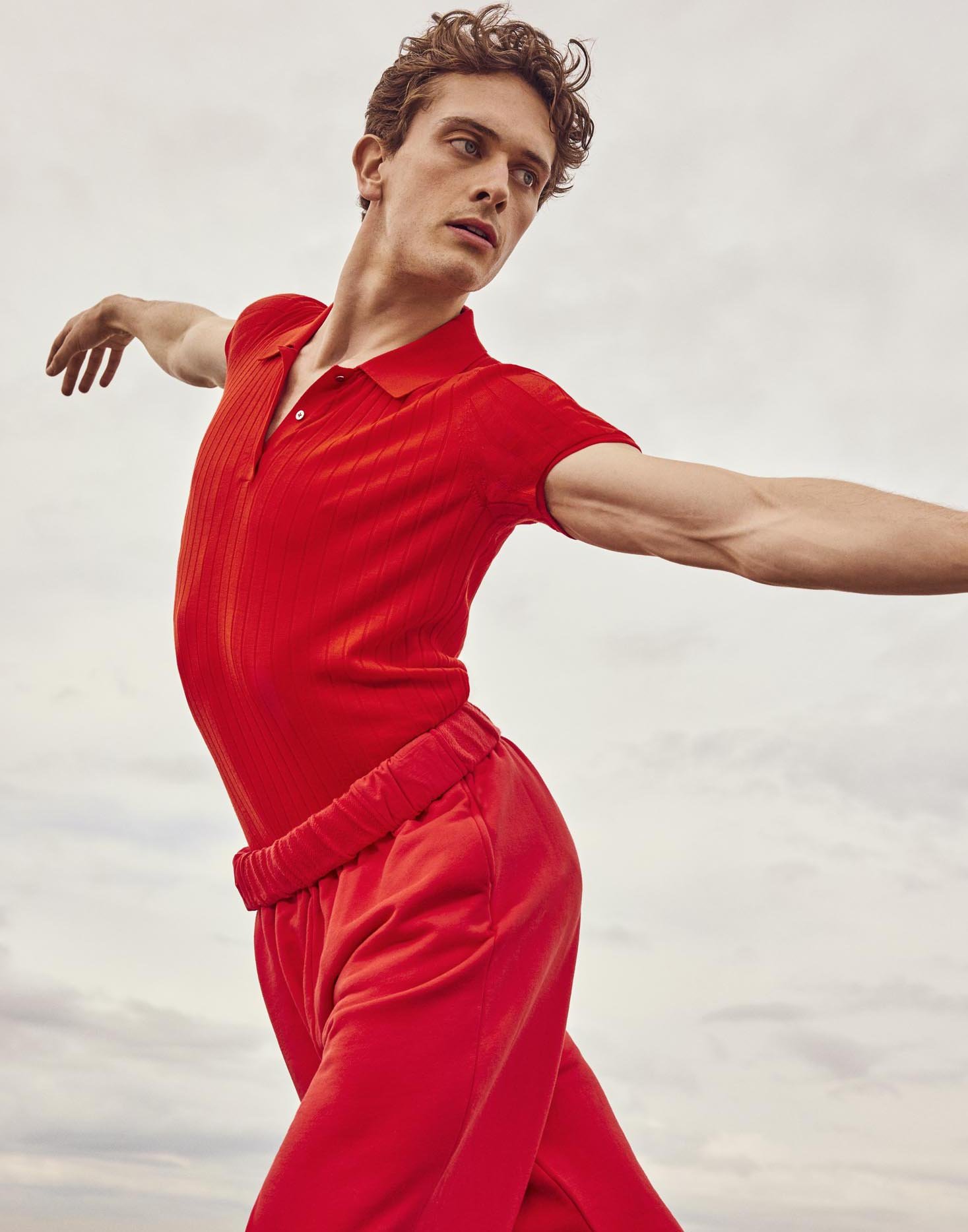 An athletic young man in bright red knitwear and pants strikes a dance pose against a soft, out-of-focus cloudy backdrop.