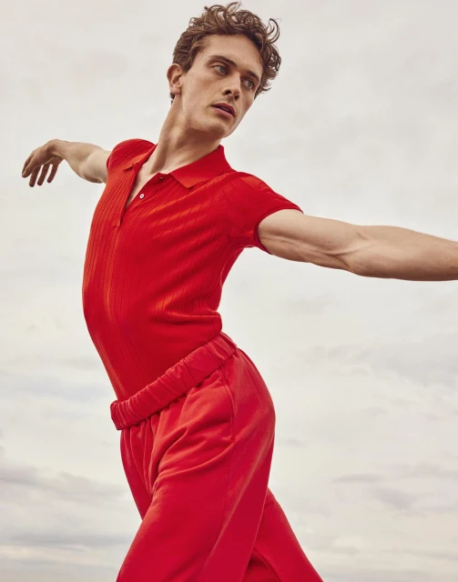 An athletic young man in bright red knitwear and pants strikes a dance pose against a soft, out-of-focus cloudy backdrop.