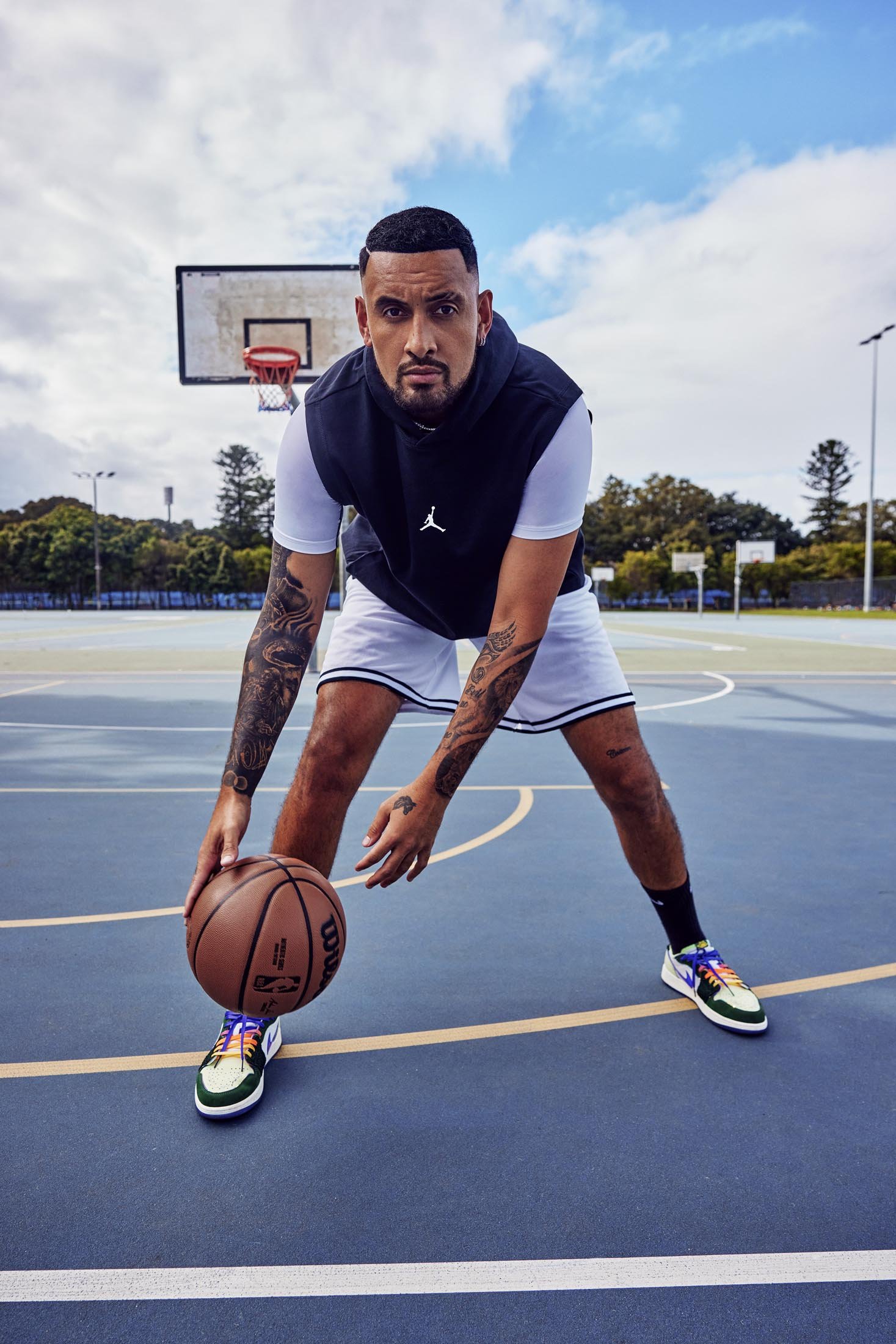 Man with tattoos dribbling basketball on outdoor court with basket, wearing black vest and white shorts, eye-level shot, daytime.