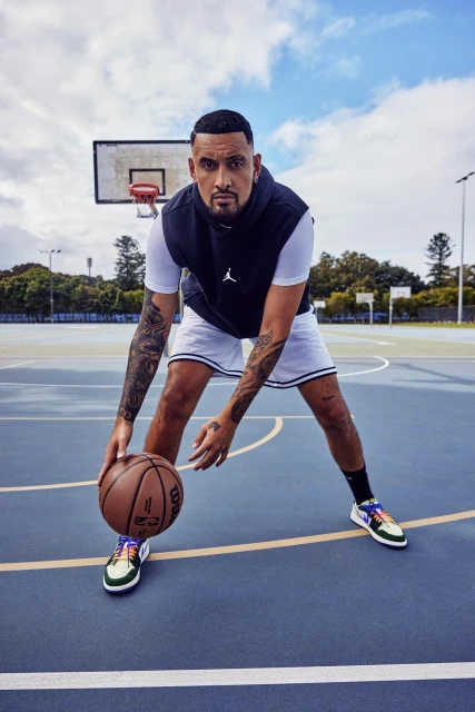 Man with tattoos dribbling basketball on outdoor court with basket, wearing black vest and white shorts, eye-level shot, daytime.